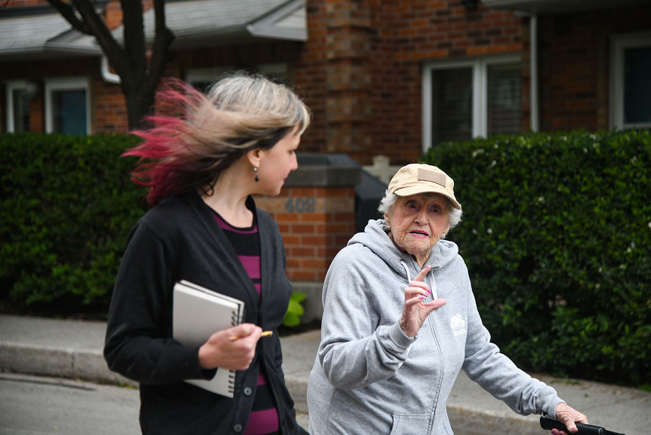 Rose Lipszyc wearing a beige hat and grey sweats while talking and walking with Miriam Libicki who is holding a notebook.