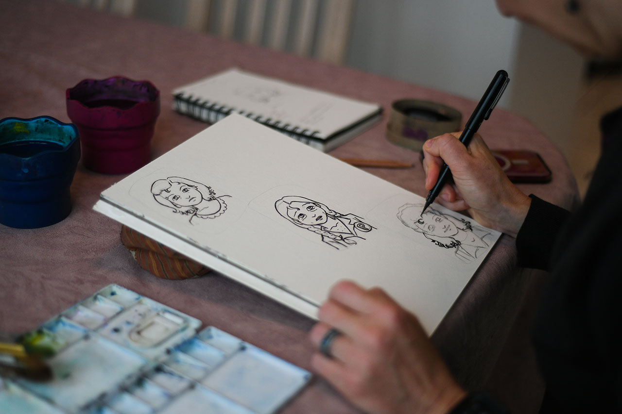 Miriam Libicki sketching some drawings on a notebook with a black pen and some watercolour paint brushes on the left.