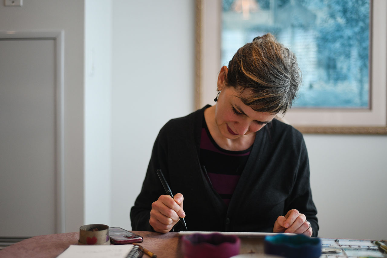 Miriam Libicki wearing a black top and sketching drawings with a black pen against a white background with a blurred blue painting.