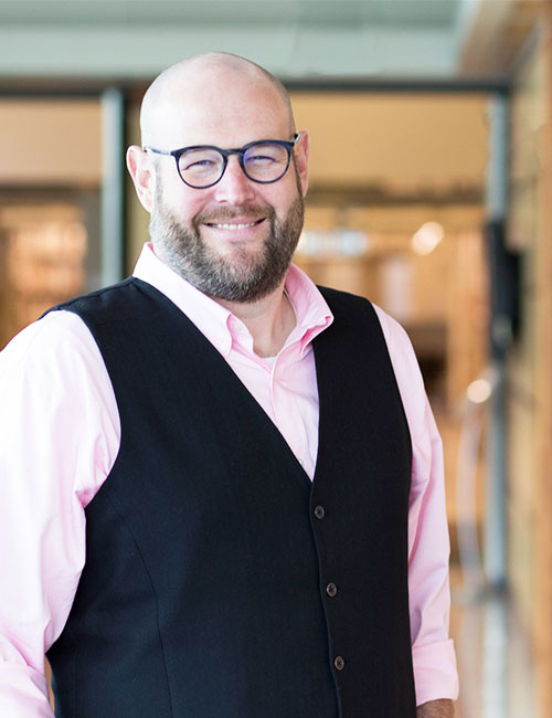 Matt Huculak wearing glasses, a pink shirt, a black vest and smiling against a blurred office space.