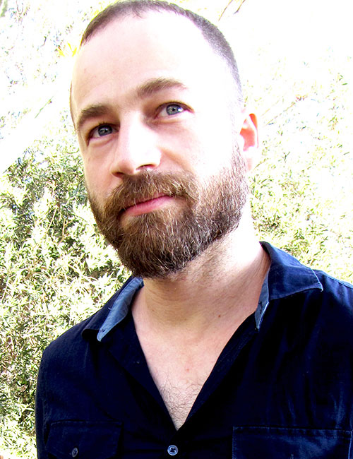 Gilad Seliktar looking at the horizon and wearing a dark blue shirt against an overexposed background with some vegetation.