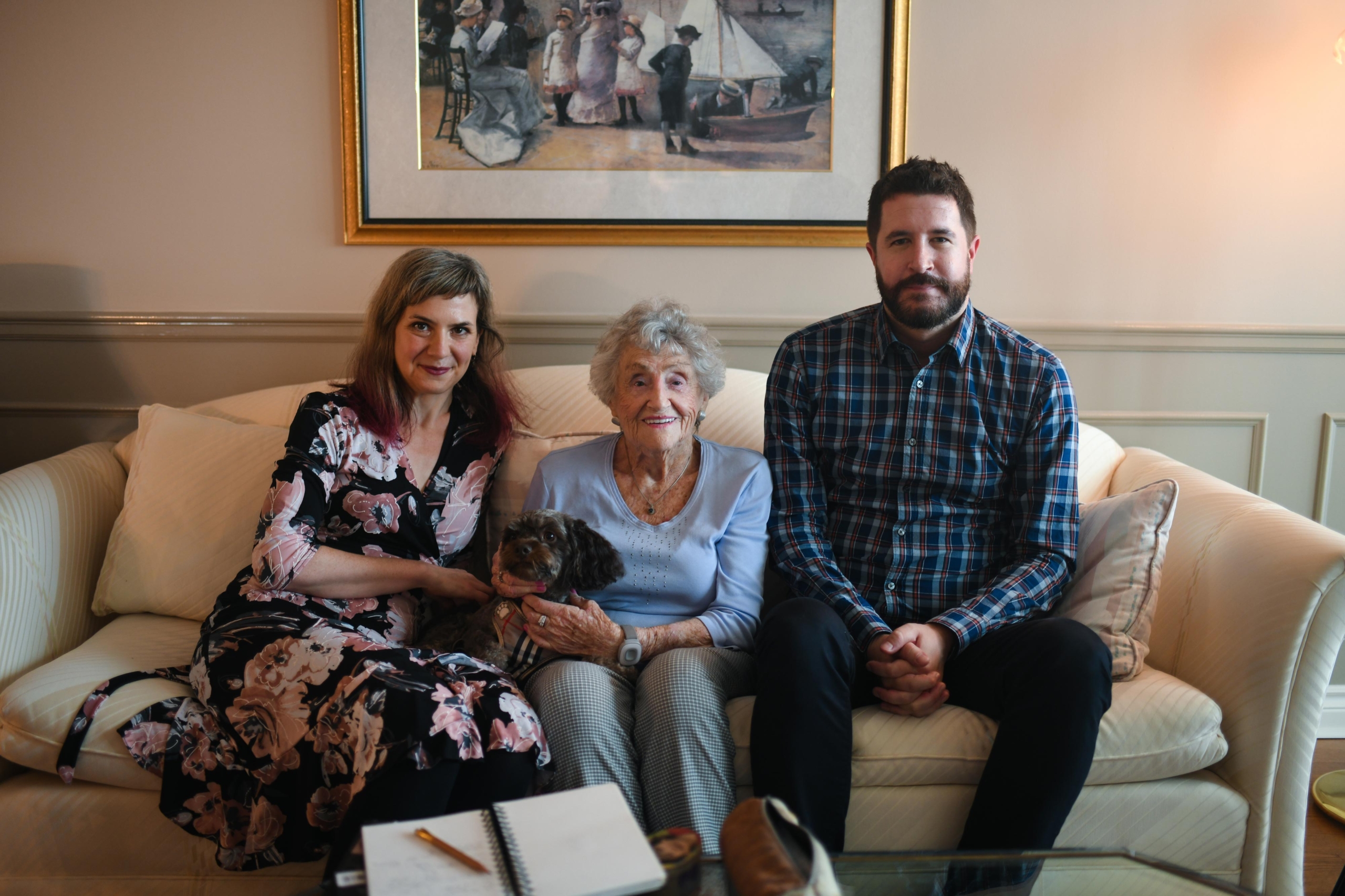 Miriam Libicki, Rose Lipszyc, a man and a dog sitting on a white couch and smiling at the camera.