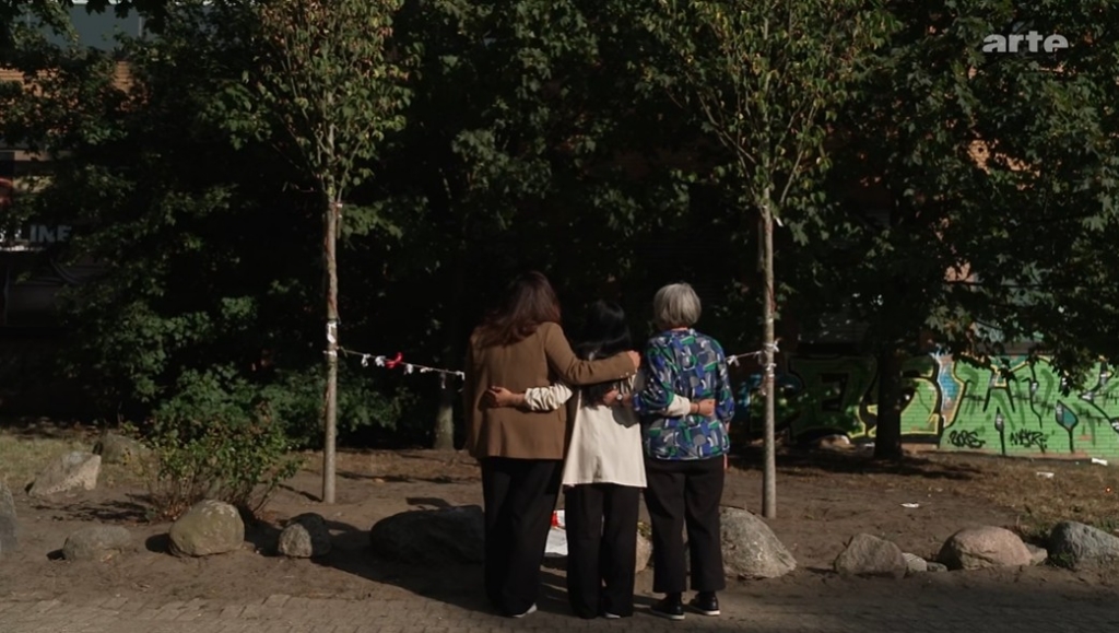 Screenshot from ARTE.TV (2026). Three women embrace outdoors surrounded by trees.